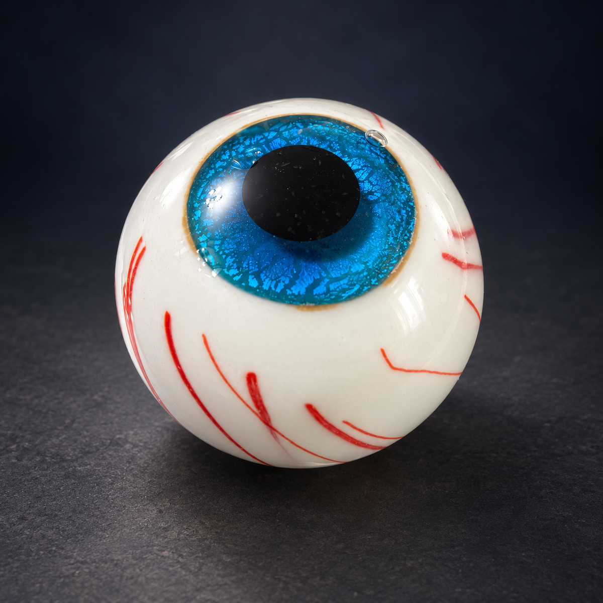 Blue eyeball glass paperweight with white body and red veins on dark navy background